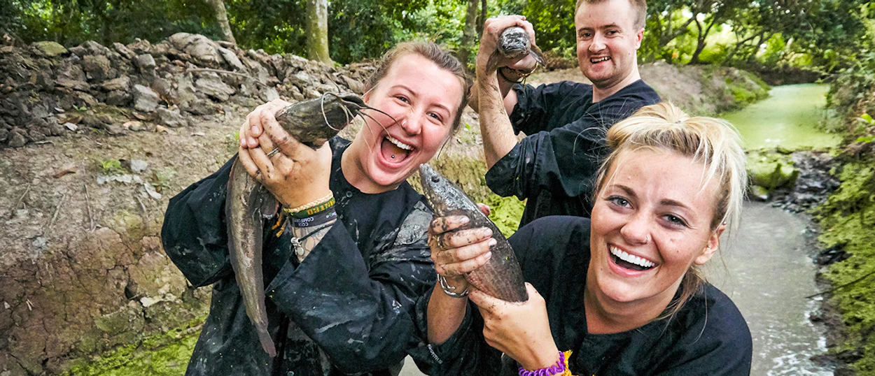 Smiling tourists covered in mud hold freshly caught fish in a rural Mekong Delta canal.