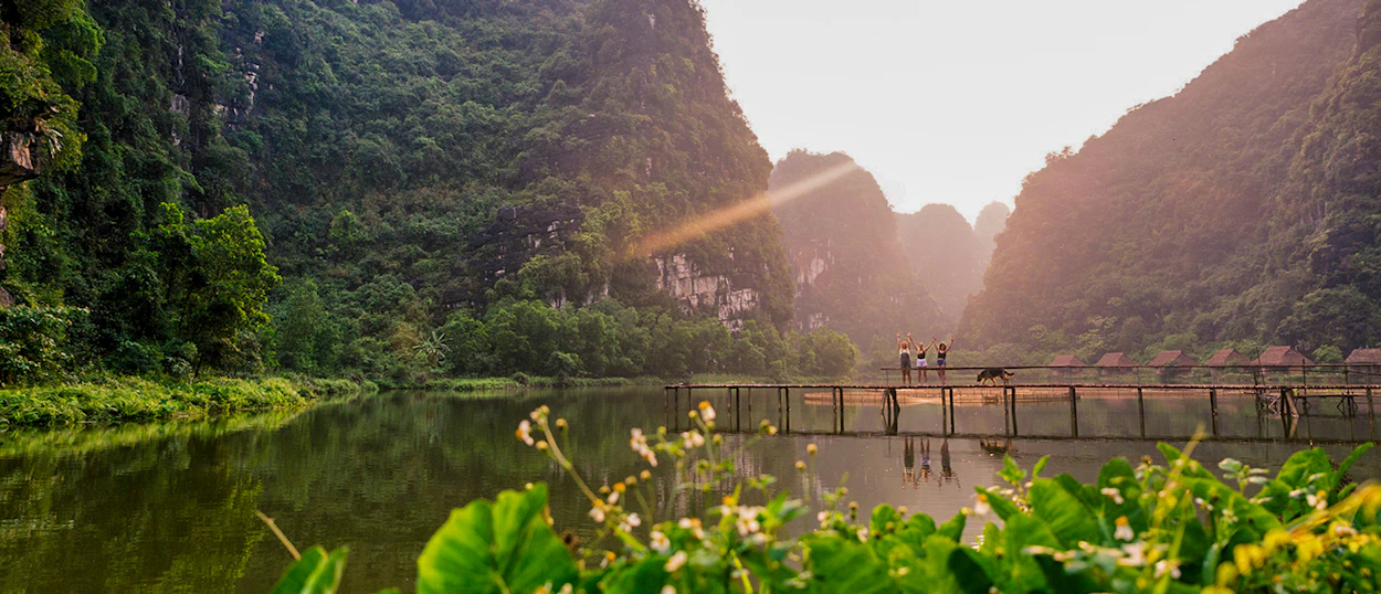 People and a water buffalo cross a wooden bridge over a calm lake at sunset in Ninh Binh.